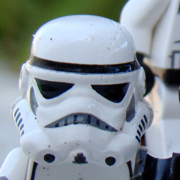 Lego Troopers Crop A close-up of a white stormtrooper helmet with distinct black markings, showcasing its iconic design. The helmet has some dirt, suggesting it has been played with or displayed outdoors, while a second stormtrooper figure is slightly visible in the background.
